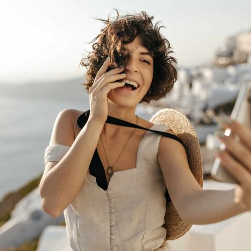 brunette-curly-woman-in-beige-dress-and-straw-hat-smiles-sincerely-and-takes-selfie-outside-in-old.jpg